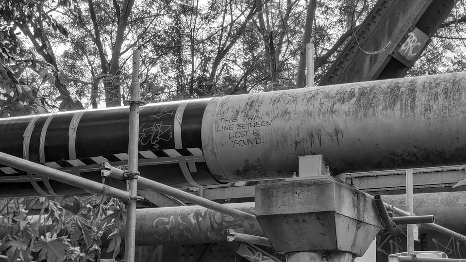 A piece of graffiti underneath a bridge in Emu Plains, New South Wales. It reads 'THE THIN LINE BETWEEN LOST & FOUND...'. A black and white filter has been applied.