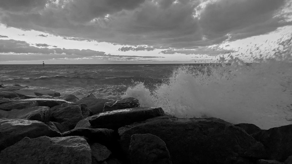During a rough tide after the sun has set, waves crash against rocks on the shore at Carrum Beach. A black and white filter has been applied.