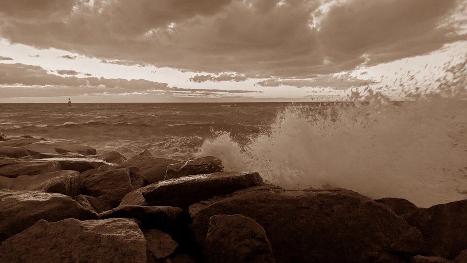 During a rough tide after the sun has set, waves crash against rocks on the shore at Carrum Beach. A sepia filter has been applied.